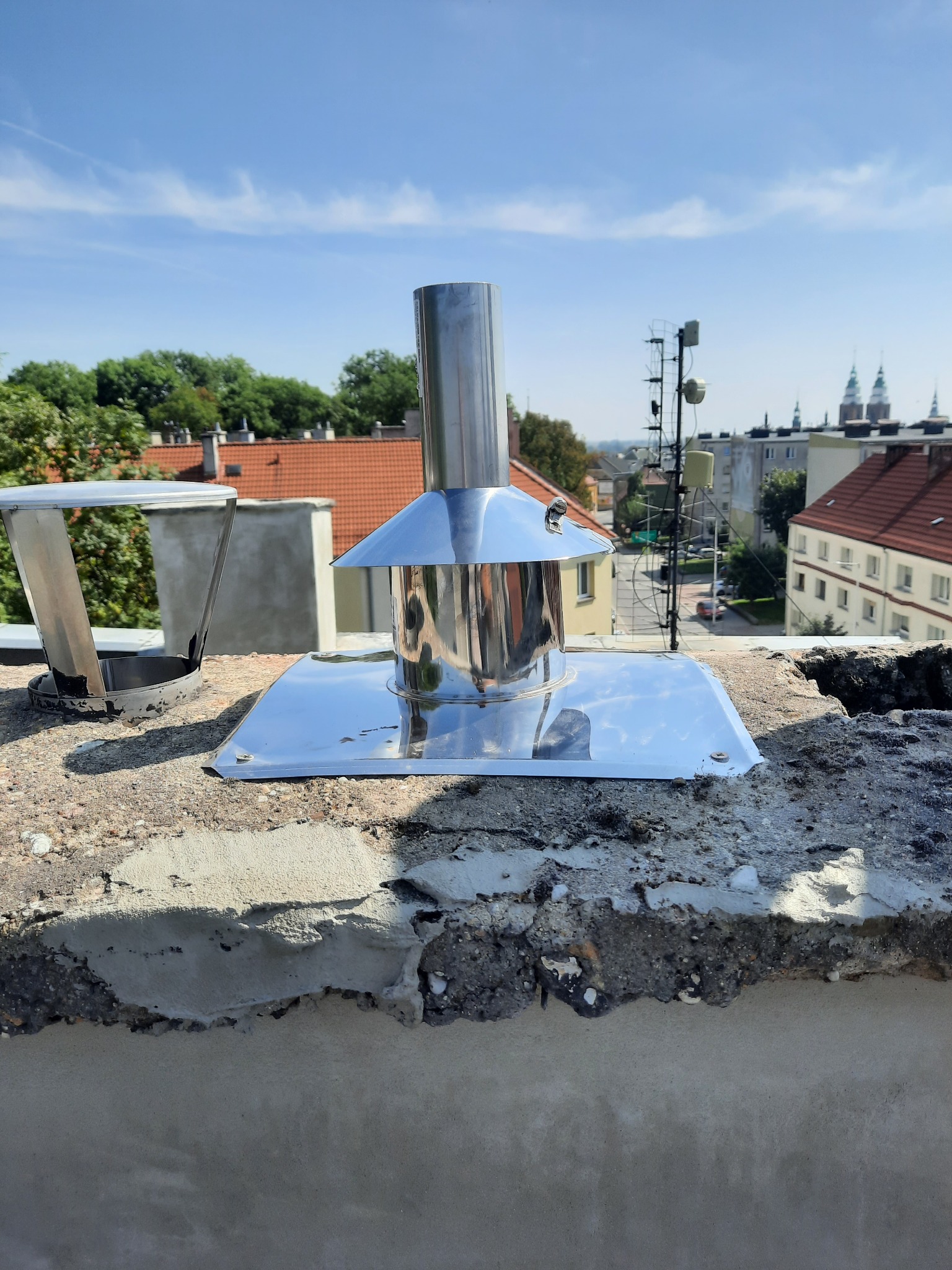 Błyszczący, nowy komin ze stali nierdzewnej zamontowany na betonowej podstawie dachu, widok na miasto w tle.
