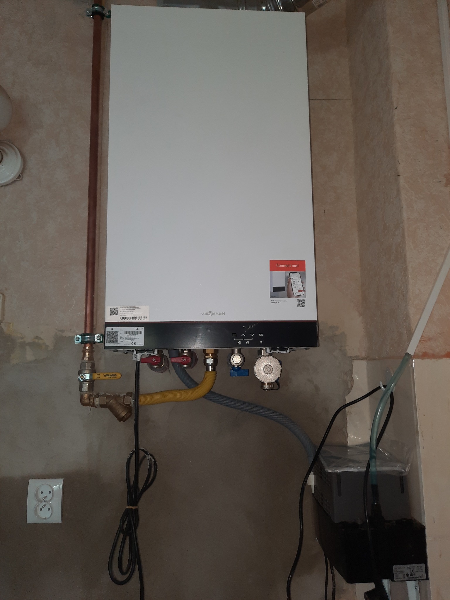 Biały piec gazowy Viessmann zamontowany na szarej ścianie, widoczne przyłącza gazowe i hydrauliczne, rura miedziana, oraz podłączenie elektryczne. Na piecu naklejka 'Connect me!'