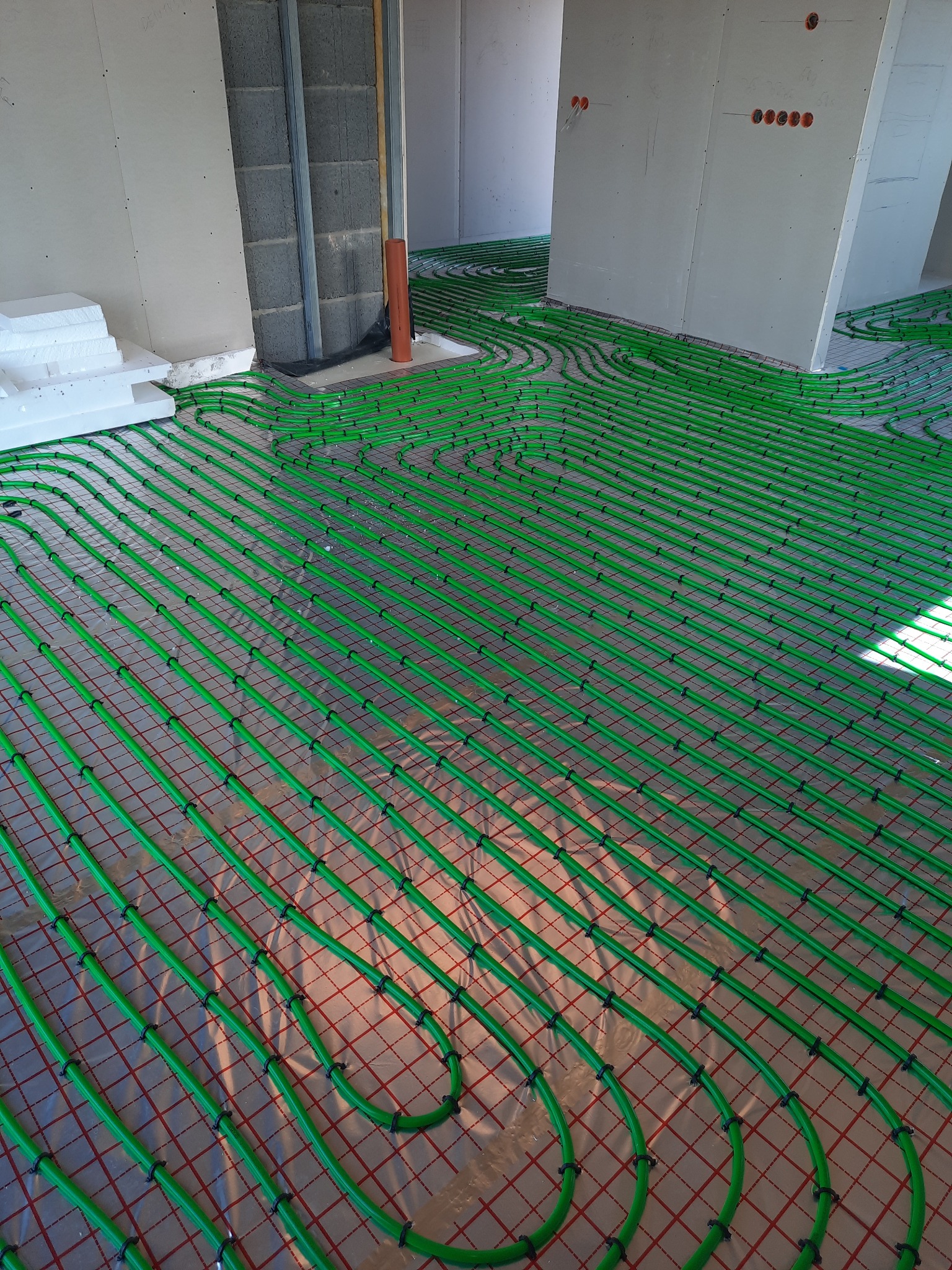 Zielone rury ogrzewania podłogowego ułożone w pętle na folii z siatką, widoczne ściany w stanie surowym i izolacja ze styropianu.