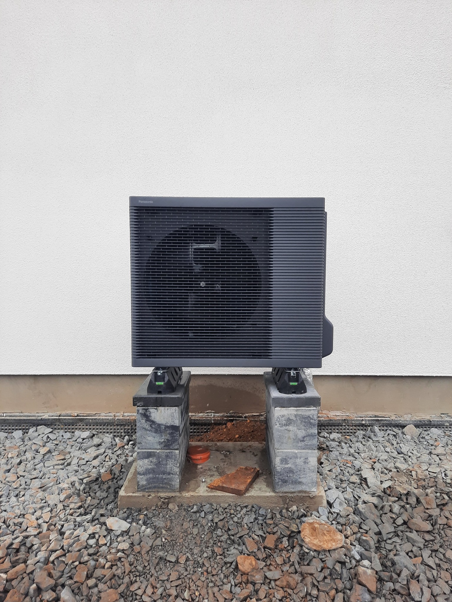 Szara pompa ciepła Panasonic zamontowana na betonowych bloczkach na tle białej ściany z kamienistym podłożem.