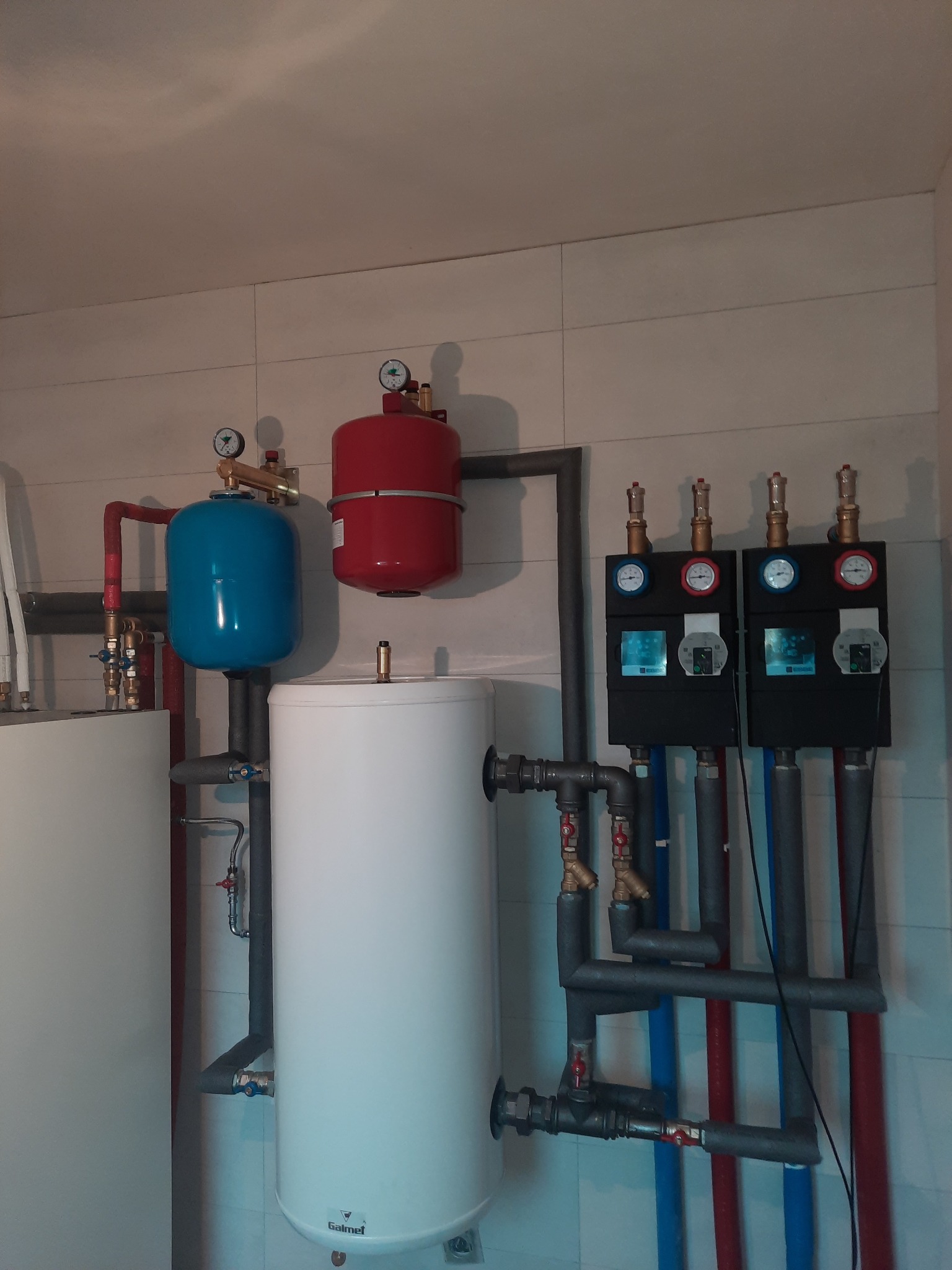 Instalacja grzewcza z dwoma zbiornikami buforowymi (biały i niebieski), czerwonym naczyniem przeponowym, rozdzielaczem z czterema obiegami grzewczymi, rurami w kolorach czerwonym i niebieskim...