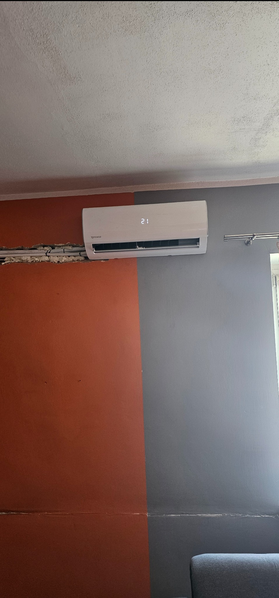 Biała klimatyzacja Rotenso na ścianie pomalowanej na pomarańczowo i szaro. Widoczne przewody w ścianie. Ujęcie z dołu, widoczny fragment sufitu i kanapy.