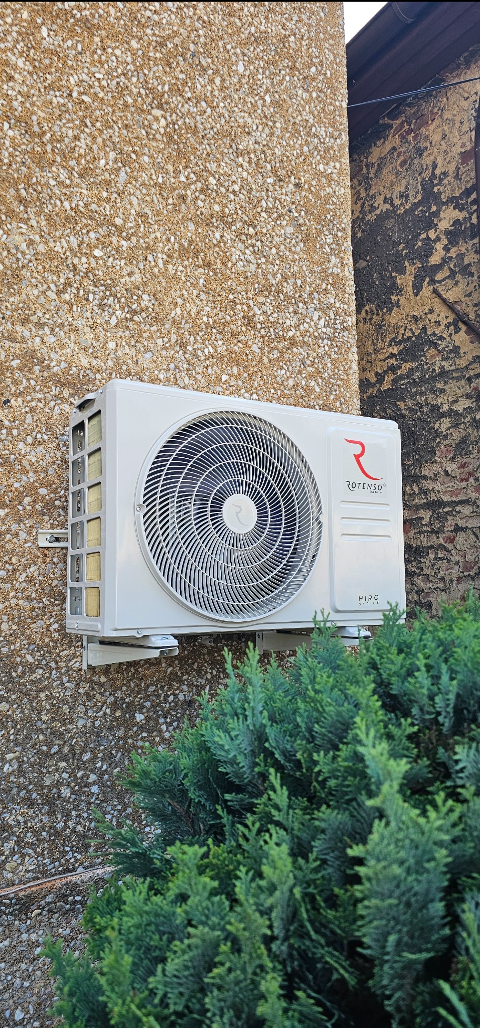 Biała pompa ciepła Rotenso, model Hiro Series, zamontowana na elewacji budynku z fakturą baranka, z zielonym krzewem u dołu kadru. Widoczne mocowania i fragment ściany sąsiedniego budynku.