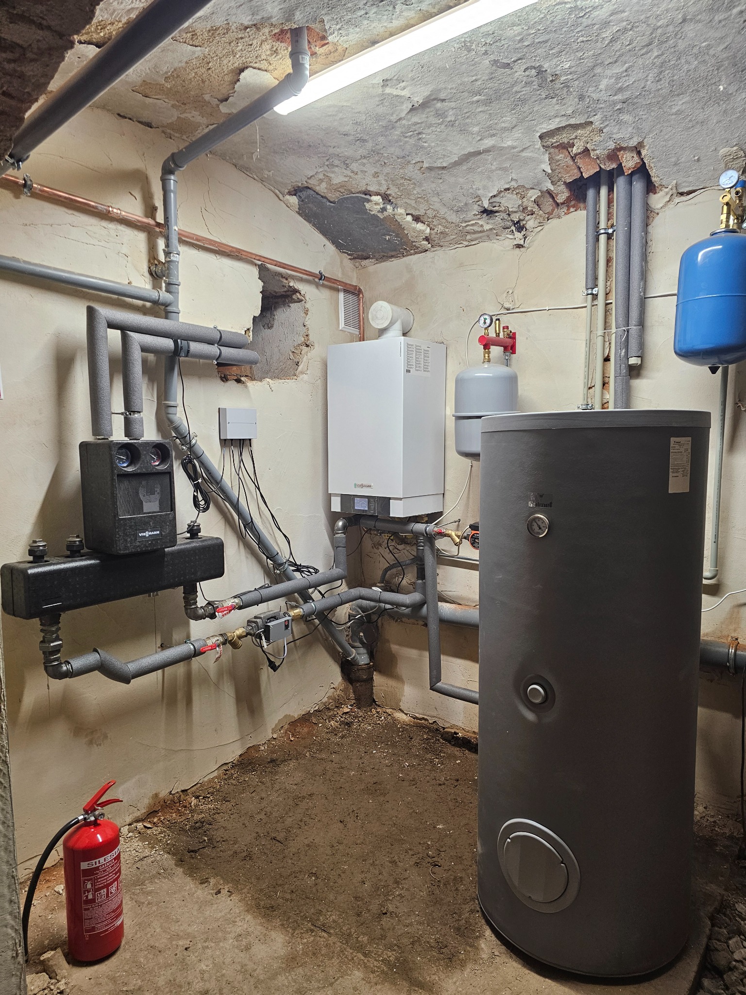 Kotłownia z piecem gazowym Viessmann, zasobnikiem Solmet i instalacją. Widoczne rury miedziane i szare, ściany z ubytkami tynku, gaśnica. Narożne ujęcie.