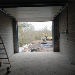 Prywatna - Wnętrze garażu z częściowo otwartą bramą segmentową, widoczny teren budowy z koparką na zewnątrz, drabina oparta o ścianę wewnątrz garażu, surowy betonowy podkład.