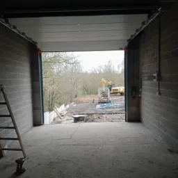 Wnętrze garażu z częściowo otwartą bramą segmentową, widoczny teren budowy z koparką na zewnątrz, drabina oparta o ścianę wewnątrz garażu, surowy betonowy podkład.