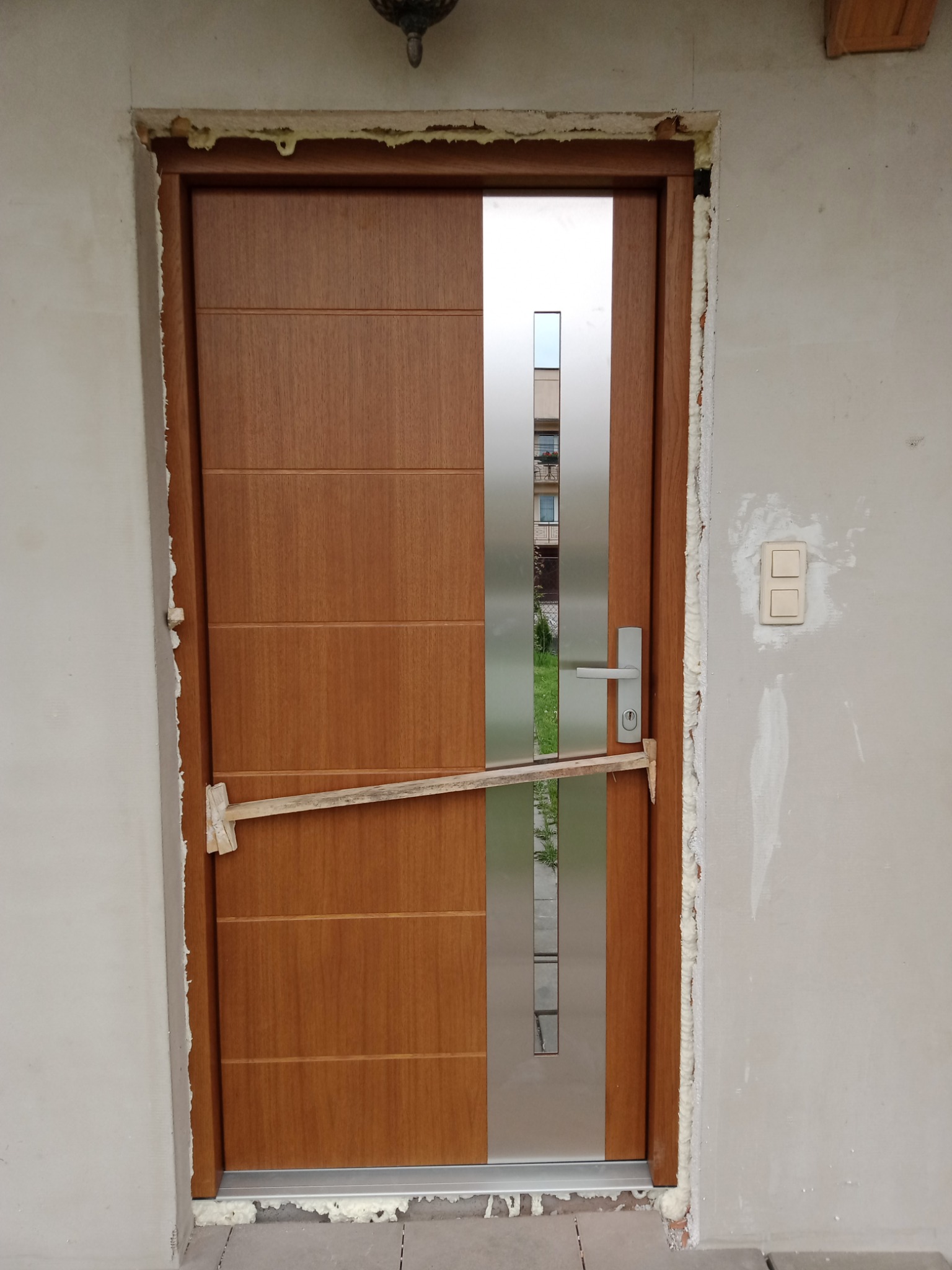 Brązowe drzwi wejściowe z pionowym, srebrnym panelem podczas montażu, zabezpieczone drewnianą rozpórką, z widoczną pianką montażową wokół ościeżnicy i niedokończonymi ścianami.