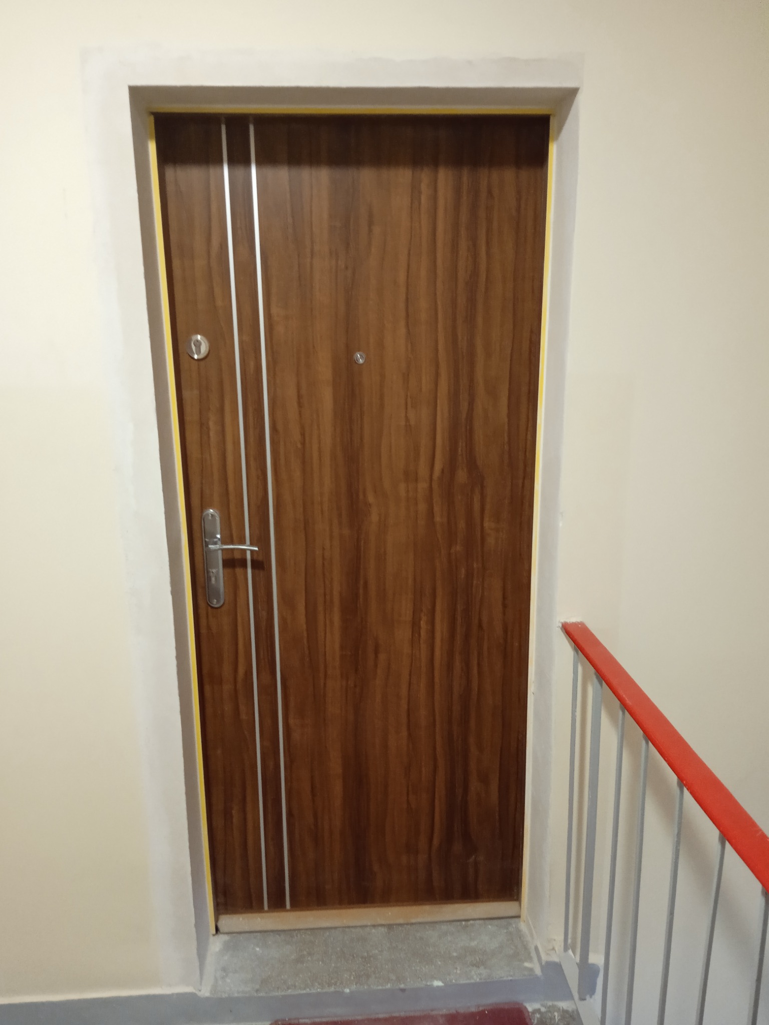 Zamontowane, brązowe drzwi wejściowe z trzema srebrnymi paskami, widoczne na klatce schodowej w Kamieniu, z pomalowaną na czerwono barierką.