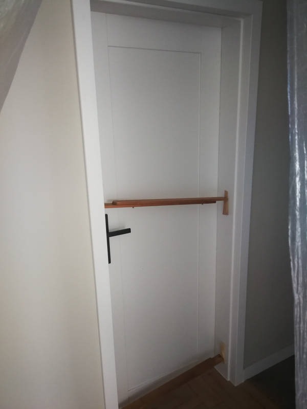 Białe, wewnętrzne drzwi z czarną klamką, zabezpieczone drewnianą poprzeczką na czas remontu, widoczne elementy podłogi i ściany.