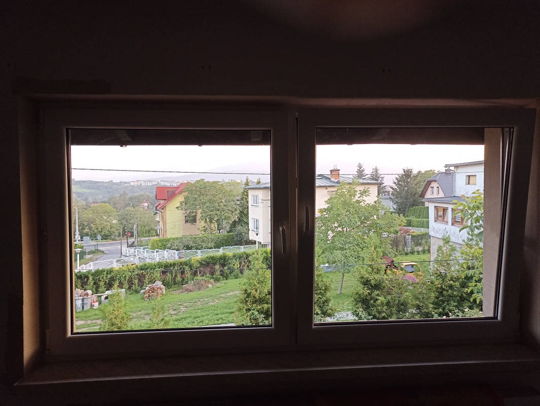 Widok z okna PCV na zielony ogród z niskim żywopłotem i zabudowę jednorodzinną w oddali, widoczne ślady prac wykończeniowych wokół ramy okiennej.