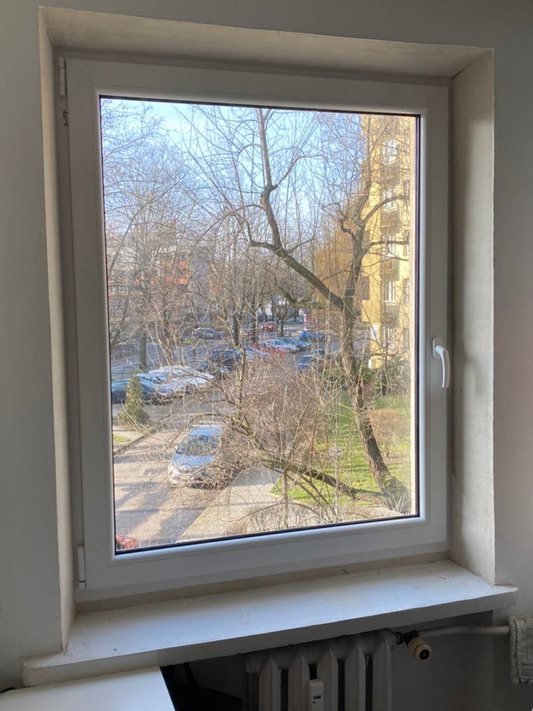 Białe okno PCV z widokiem na drzewa bez liści, zaparkowane samochody i budynek w tle; widoczny fragment grzejnika pod parapetem.