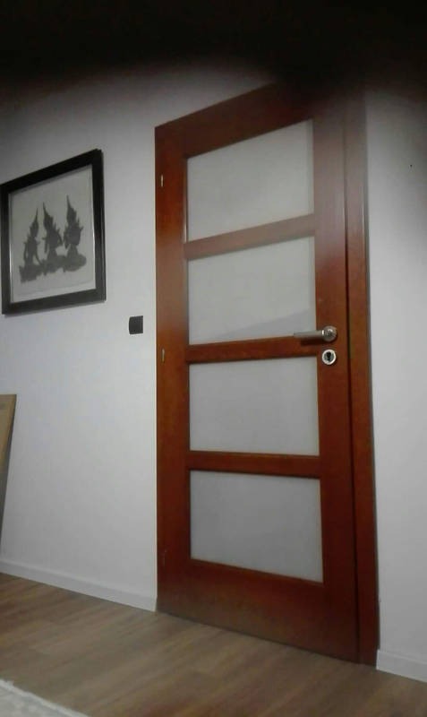 Brązowe drzwi wewnętrzne z czterema panelami z matowego szkła, zamontowane w białej ścianie, widoczne z boku, z czarną ramką z obrazem na ścianie.