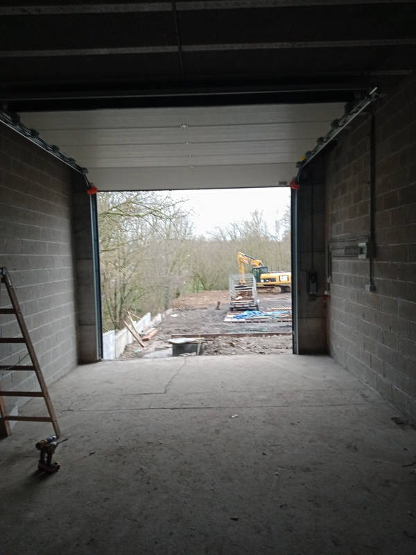 Wnętrze garażu z częściowo otwartą bramą segmentową, widoczny teren budowy z koparką na zewnątrz, drabina oparta o ścianę wewnątrz garażu, surowy betonowy podkład.