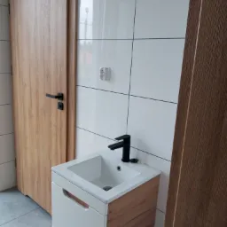 Nowoczesna łazienka z białą umywalką nablatową na szafce z drewnianymi akcentami, czarną armaturą i białymi płytkami na ścianie, obok drewniane drzwi z czarną klamką.