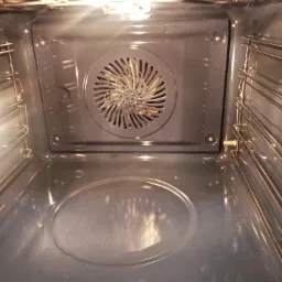 Wnętrze piekarnika elektrycznego z widocznymi prowadnicami na blachy i wentylatorem termoobiegu, po czyszczeniu, z delikatnymi smugami światła.