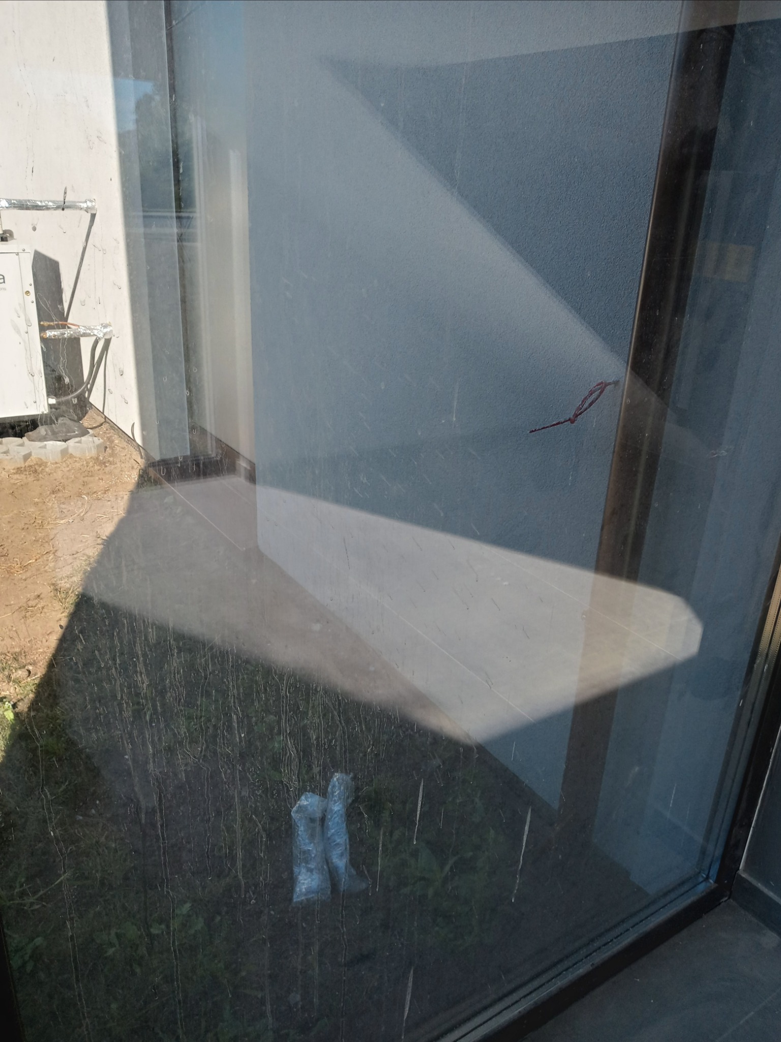 Brudna szyba okienna z widocznymi smugami i zabrudzeniami, przez którą widać fragment trawnika, elewację budynku z klimatyzatorem i dwa worki foliowe leżące na ziemi.