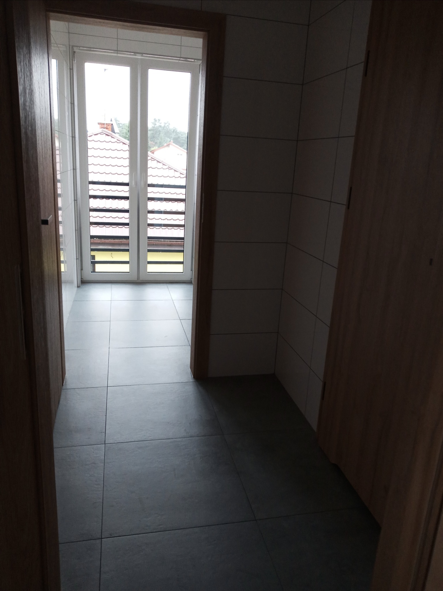 Wnętrze nowo wykończonej łazienki z szarymi płytkami podłogowymi, widok z korytarza przez drewniane futryny drzwiowe na okno z widokiem na dom z czerwonym dachem.