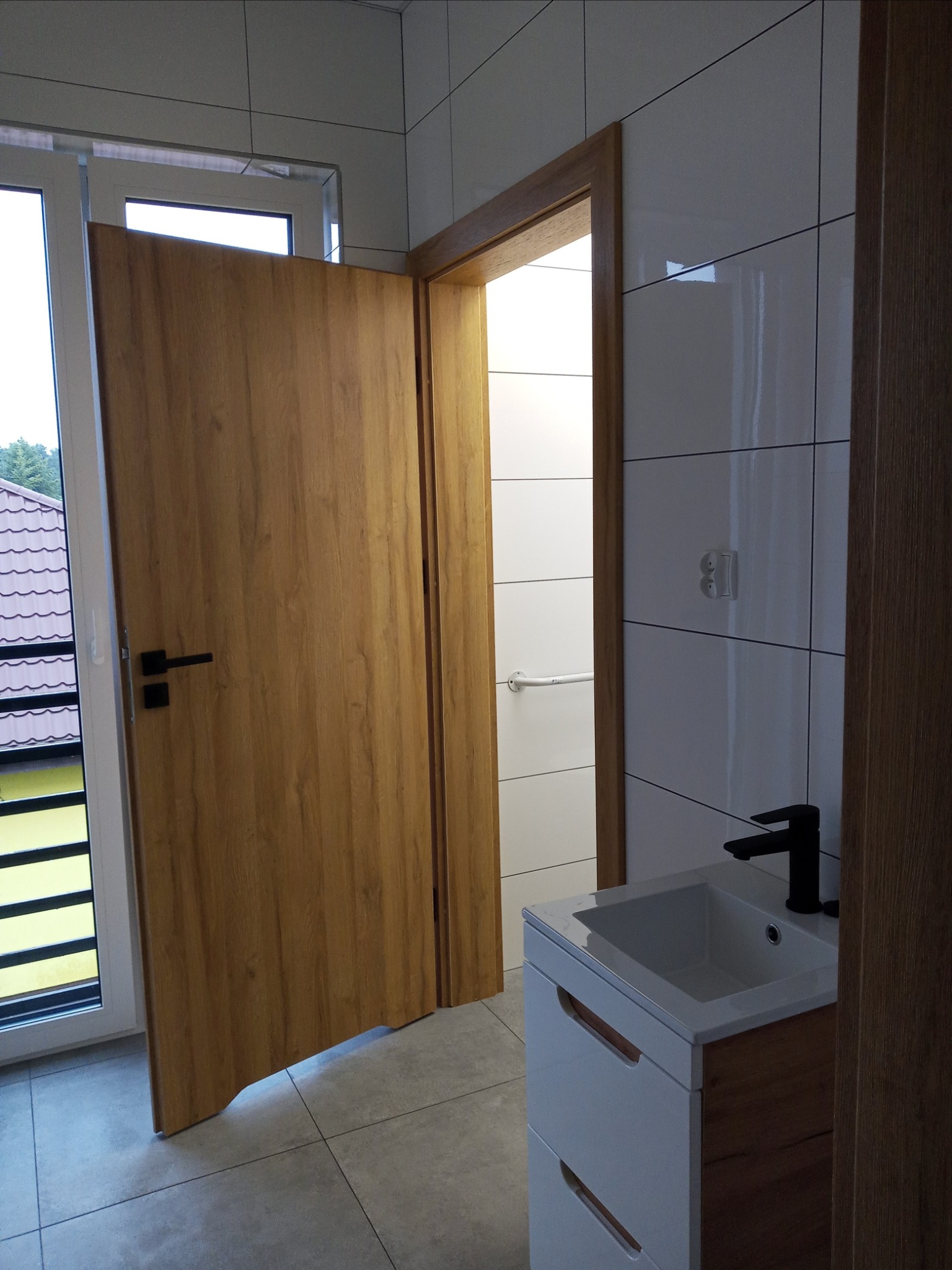 Nowoczesna łazienka z drewnianymi akcentami: otwarta drewniana drzwi z czarną klamką, białe płytki ścienne z ciemnymi fugami, umywalka z szafką w kolorze białym i drewna z czarną baterią, szare...