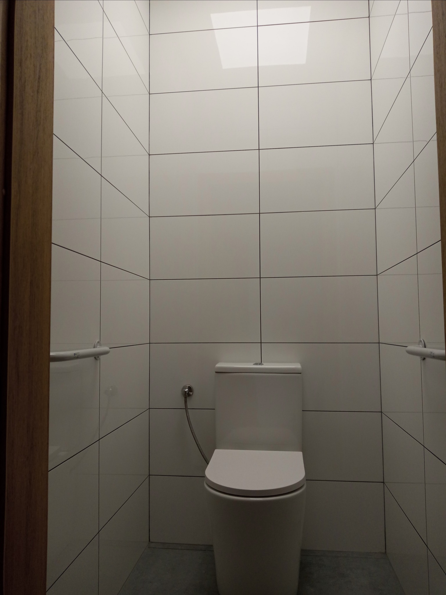 Toaleta w małej, wykafelkowanej łazience z uchwytami dla niepełnosprawnych, widok zza futryny drzwi.