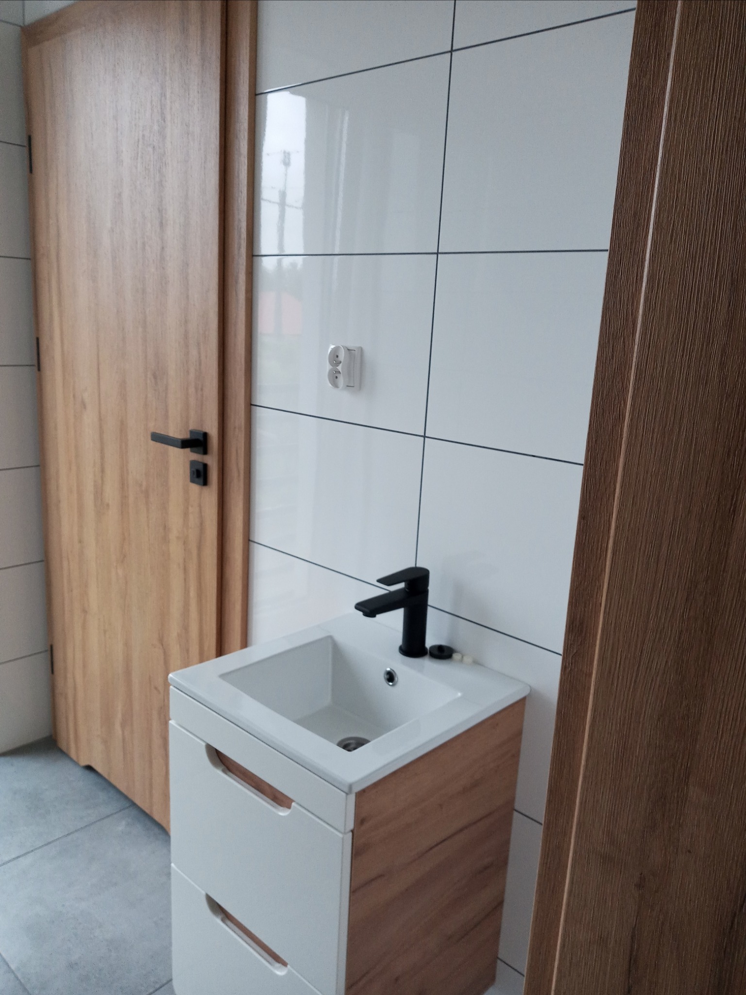 Nowoczesna łazienka z białą umywalką nablatową na szafce z drewnianymi akcentami, czarną armaturą i białymi płytkami na ścianie, obok drewniane drzwi z czarną klamką.