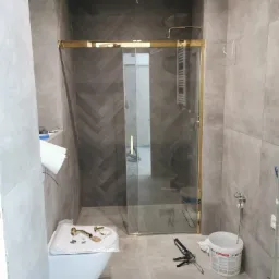 Montaż kabiny prysznicowej z złotymi profilami w łazience wykończonej szarą płytką, widoczne narzędzia i elementy instalacyjne, częściowo zamontowana toaleta.