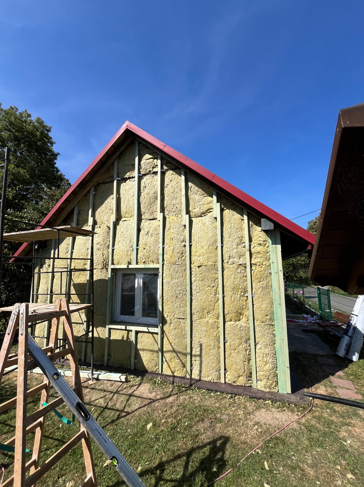 Ocieplanie ścian zewnętrznych budynku wełną mineralną, widoczne rusztowania i poziomica. Czerwony dach i błękitne niebo w tle. Prace remontowe w toku.