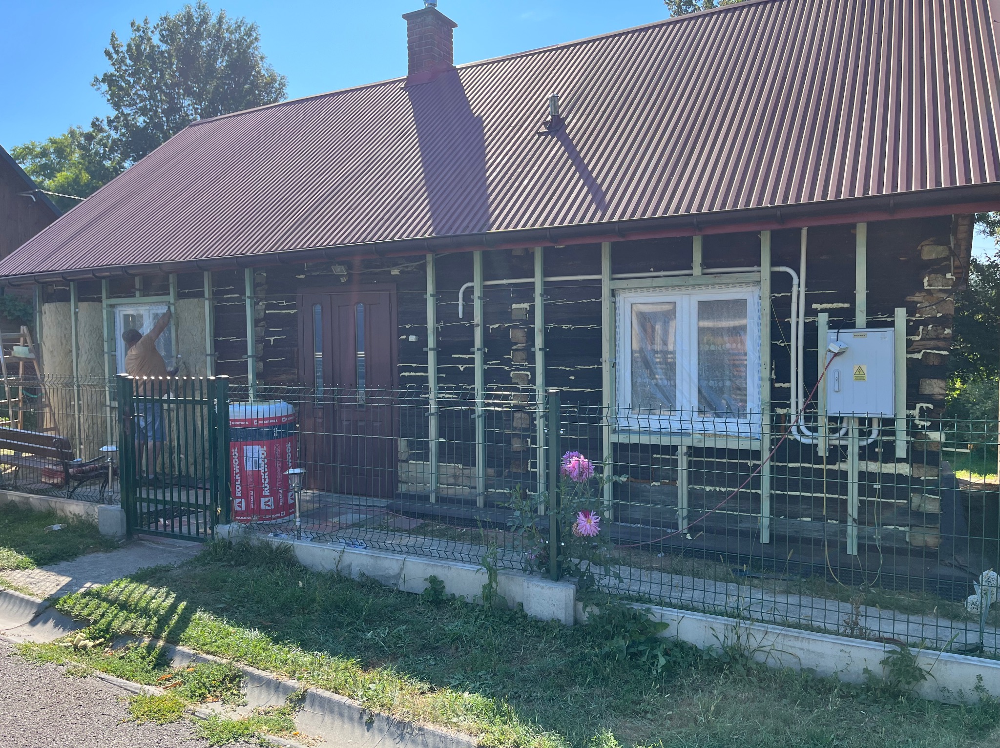 Ocieplanie drewnianego domu w Rzeszowie. Widoczne rusztowanie, puszki z wełną mineralną Rockwool i pracownik w trakcie montażu izolacji. Prace remontowe.
