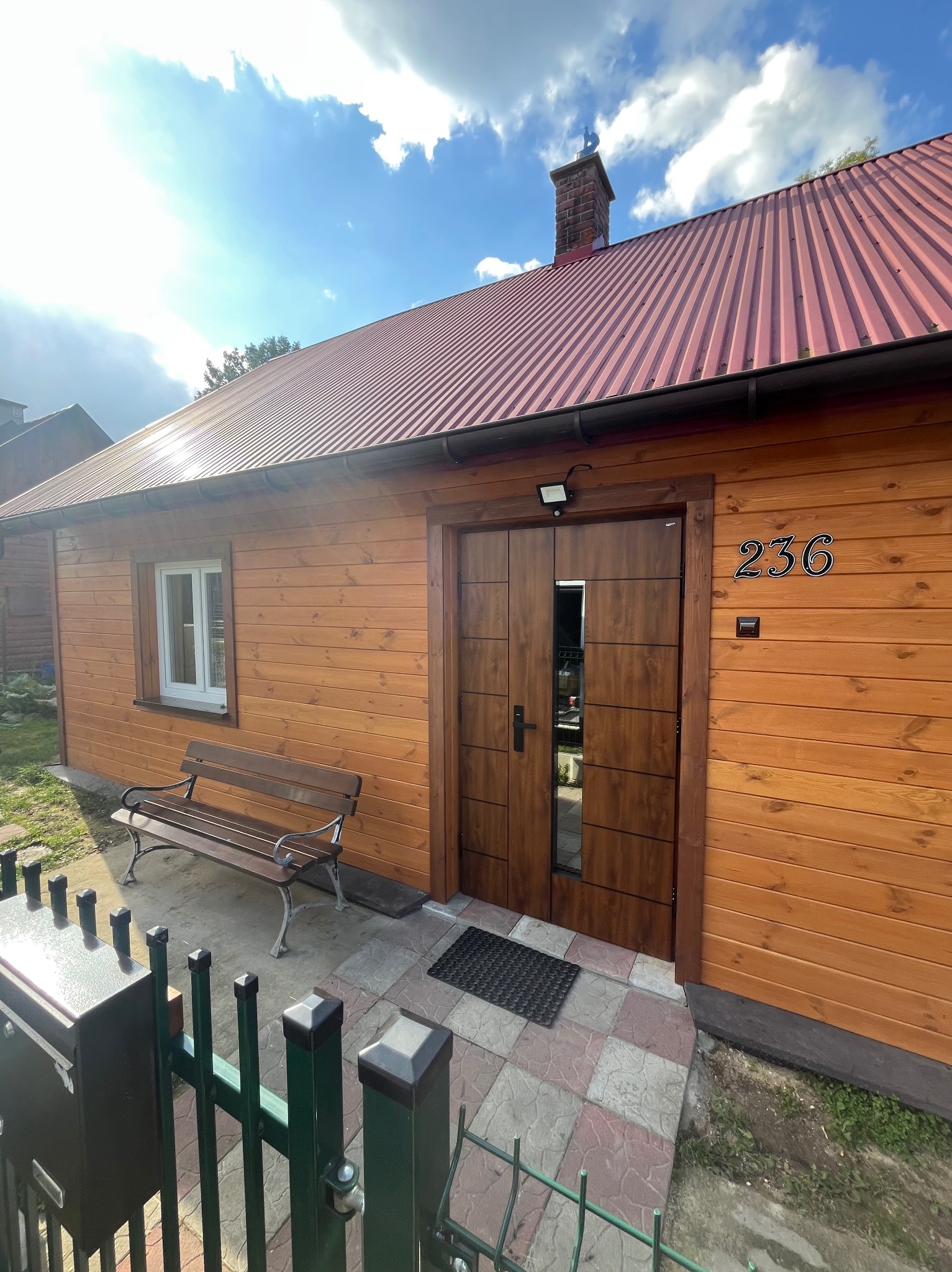 Drewniany dom z czerwonym dachem, numerem 236 i ławką przed wejściem. Widoczne nowe okna i drzwi. Całość w jasnym, słonecznym oświetleniu.