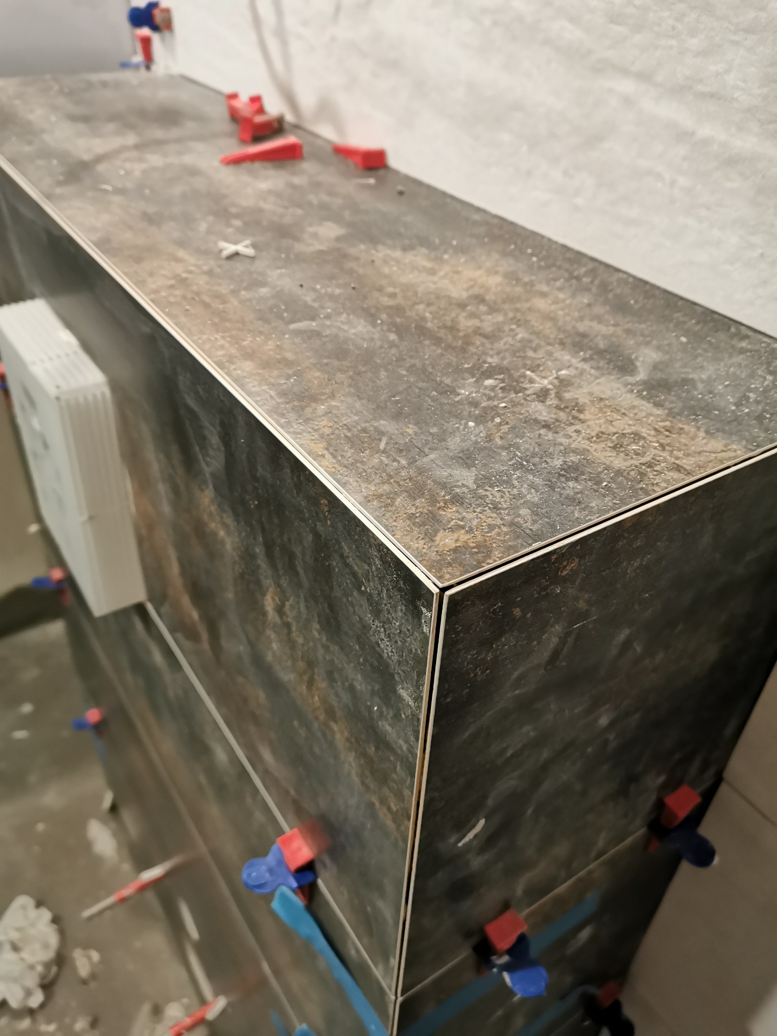 Układanie ciemnych, rustykalnych płytek na ścianie z użyciem systemu poziomowania płytek (klipsy i kliny). Widoczny biały odpowietrznik. Nieukończona realizacja.