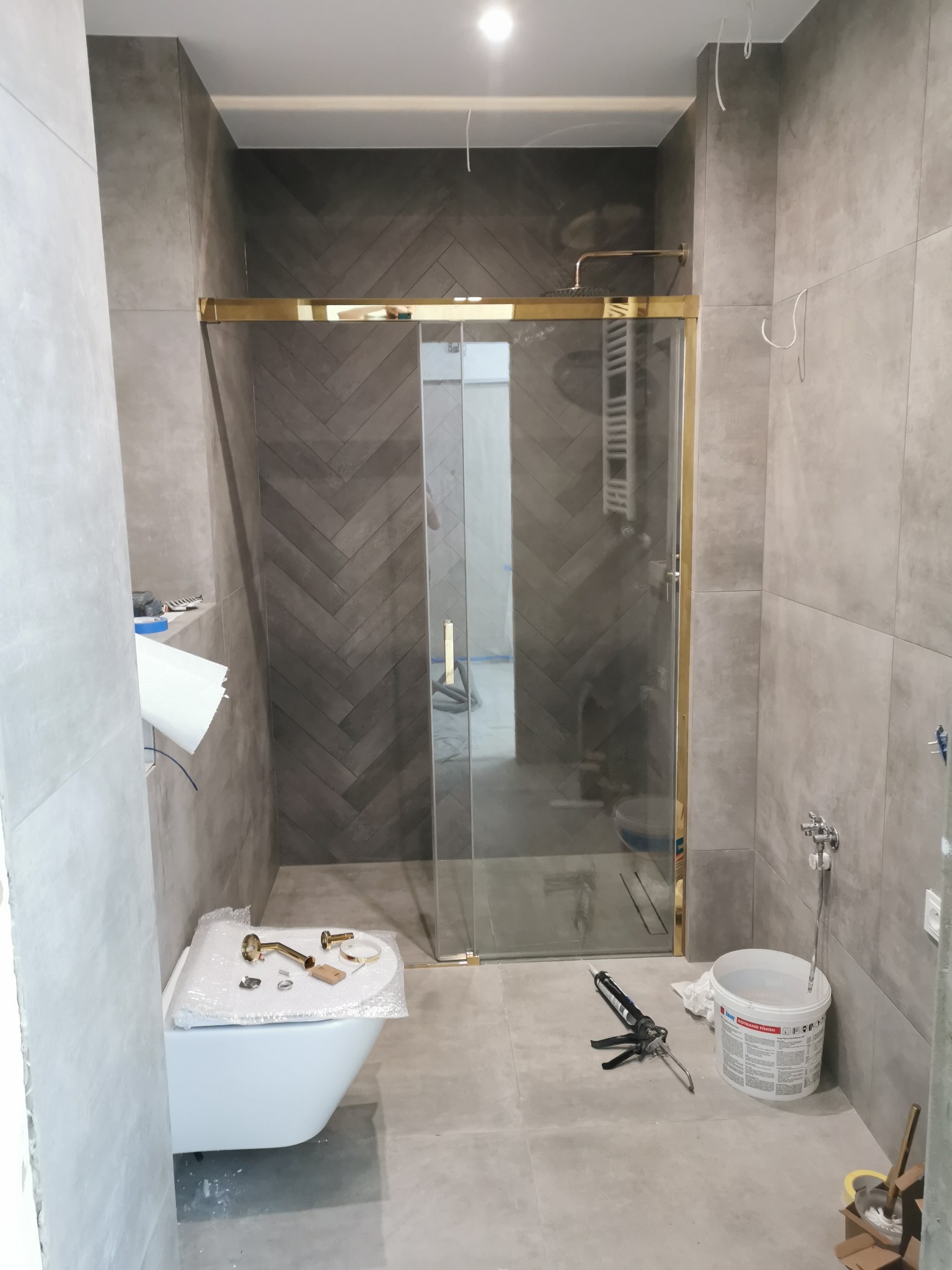 Montaż kabiny prysznicowej z złotymi profilami w łazience wykończonej szarą płytką, widoczne narzędzia i elementy instalacyjne, częściowo zamontowana toaleta.