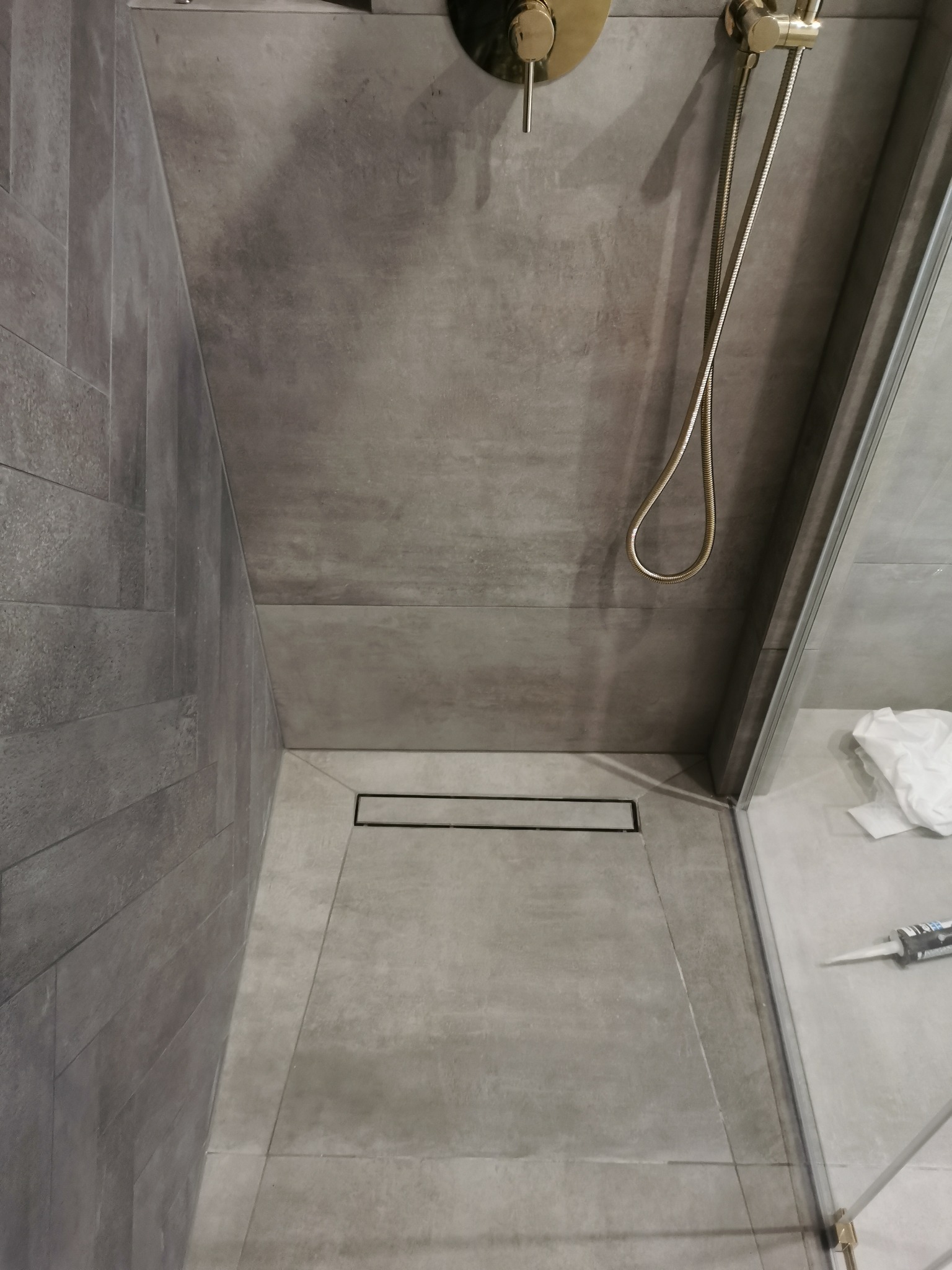 Nowoczesna kabina prysznicowa z odpływem liniowym w podłodze, wykończona szarymi płytkami imitującymi beton, złota armatura, widoczny silikon i ściereczka.
