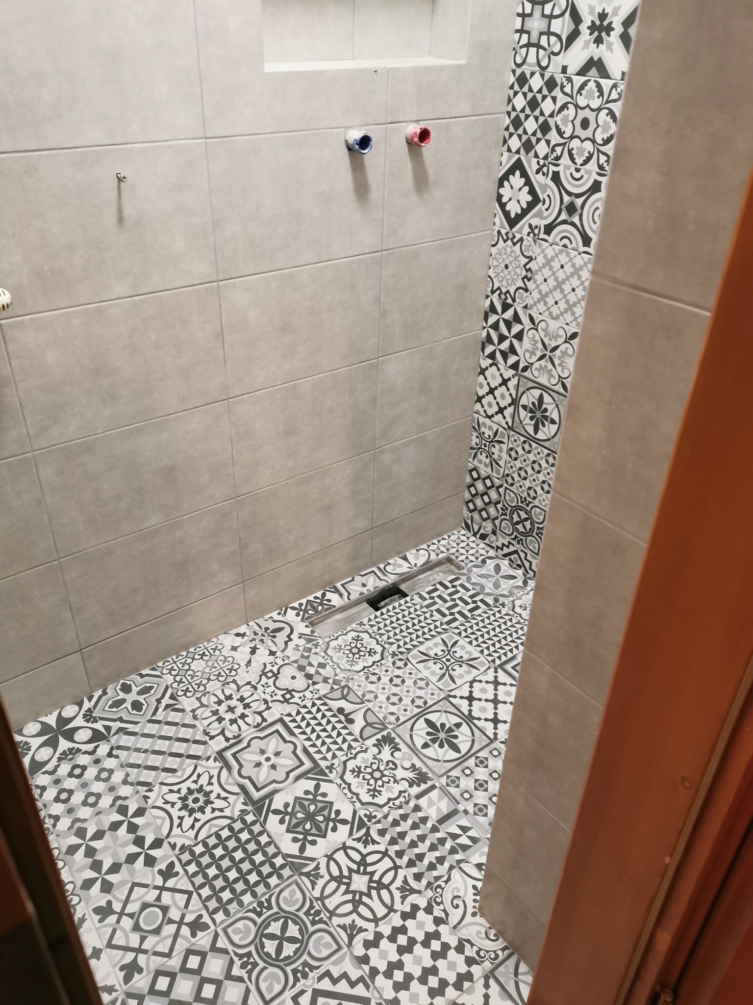 Nowoczesna łazienka w trakcie wykańczania, widoczne szare płytki ścienne, wzorzysta podłoga z odpływem liniowym oraz wystające rury instalacji hydraulicznej.