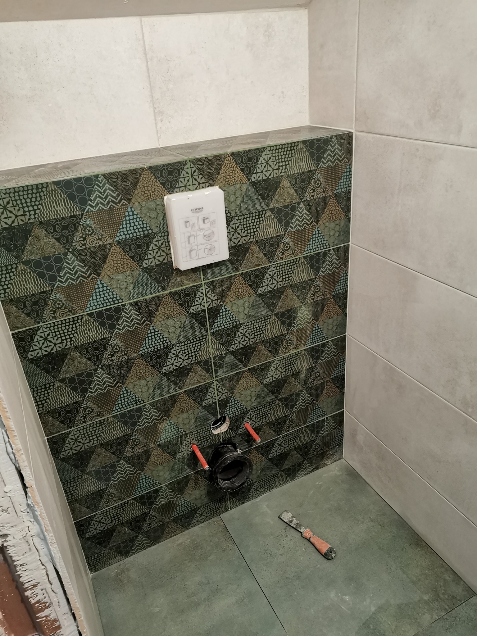 Montaż podtynkowego stelaża WC na tle ściany wyłożonej wzorzystymi płytkami, widoczne rury doprowadzające wodę oraz szpachelka na podłodze.