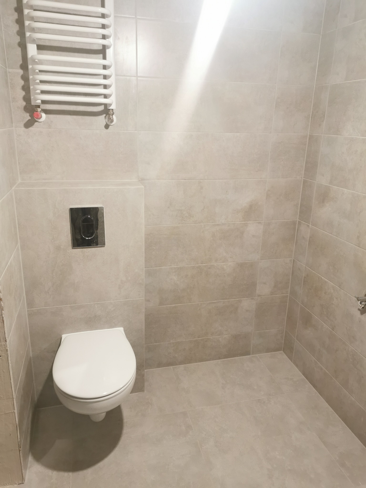 Nowoczesna, beżowa łazienka z podwieszaną toaletą, chromowanym przyciskiem spłukującym i grzejnikiem drabinkowym.