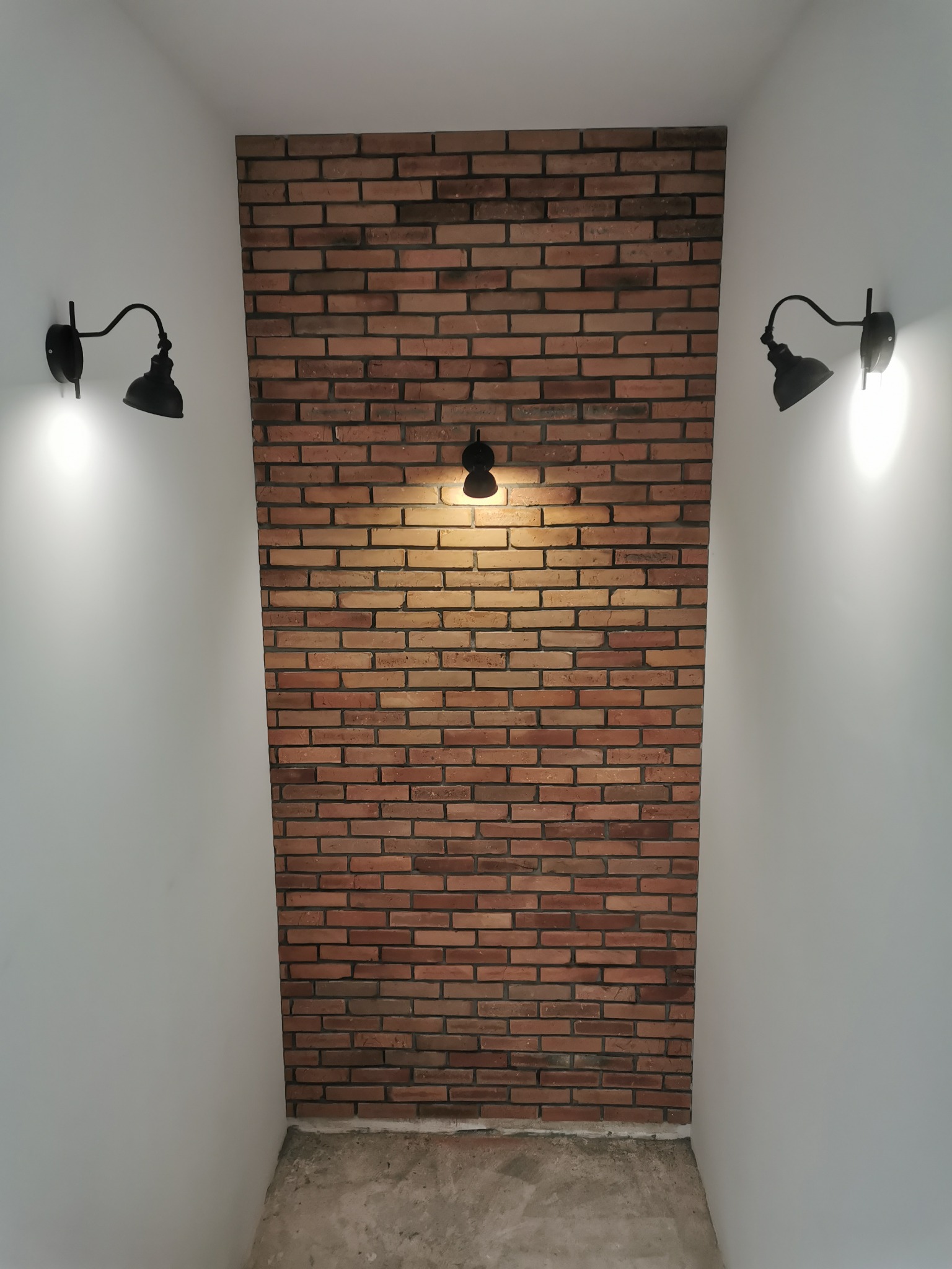 Wnętrze z dekoracyjną ścianą z cegły, oświetloną trzema czarnymi lampami ściennymi w stylu industrialnym, na tle białych ścian i surowej betonowej podłogi.