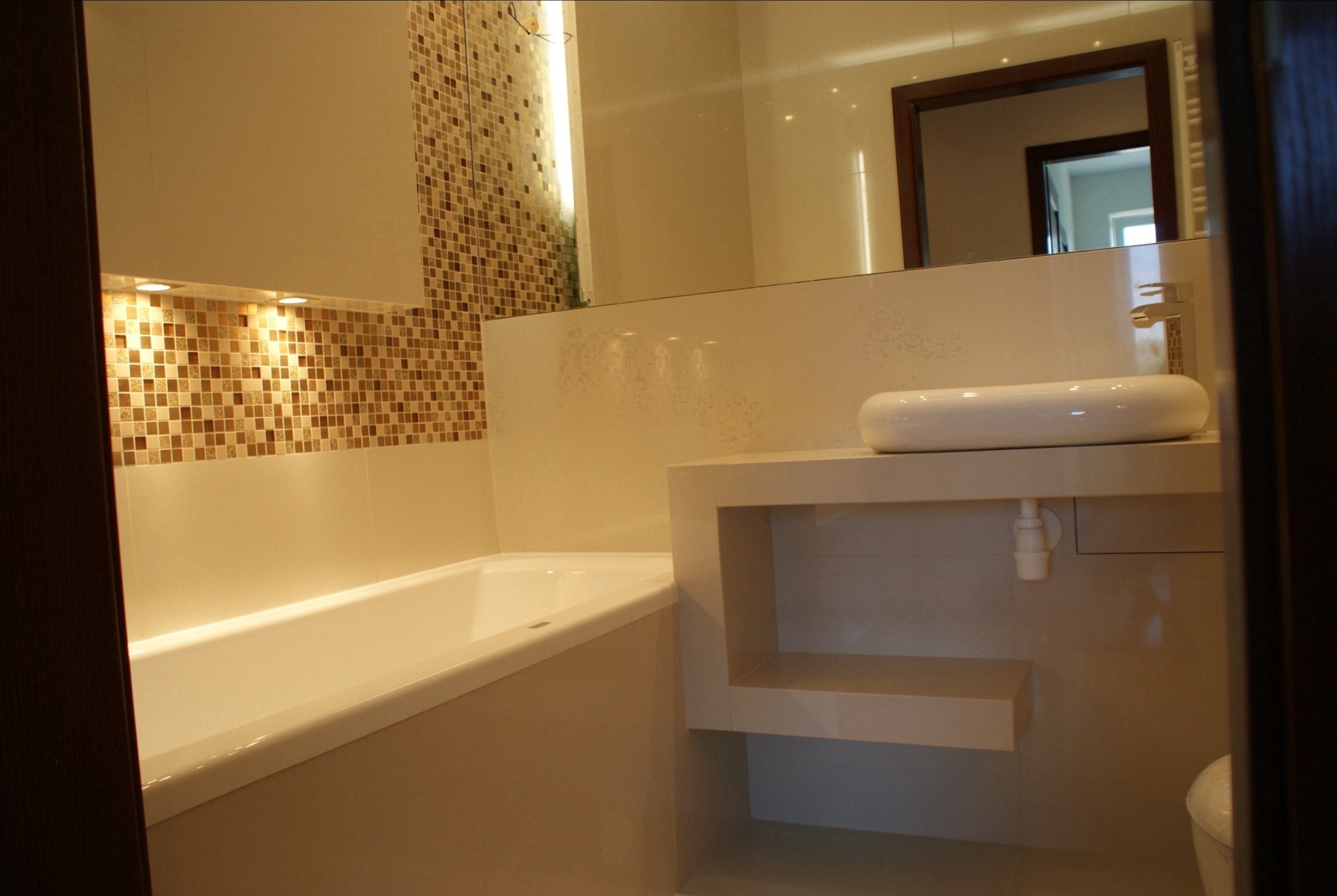 Nowoczesna łazienka z wanną, umywalką nablatową na szafce z półkami i mozaiką na ścianie, odbicie lustra w tle.