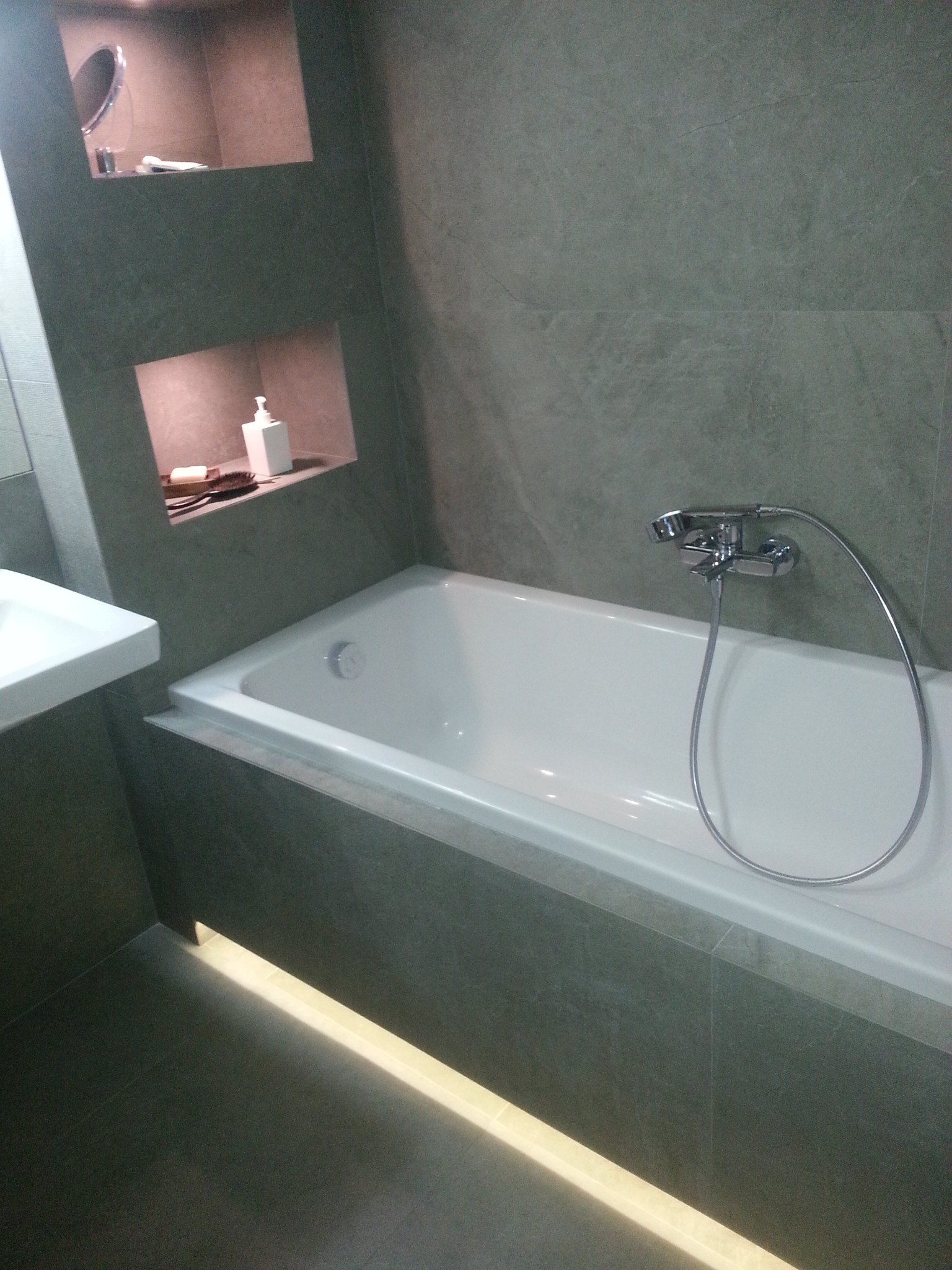 Nowoczesna łazienka z białą wanną, szarą glazurą, wnękami z kosmetykami i podświetleniem pod wanną.