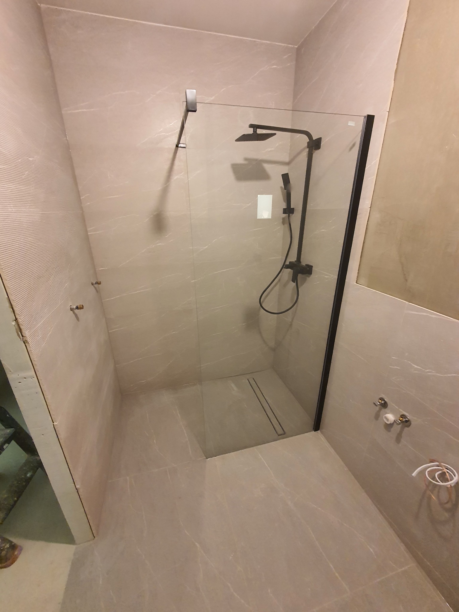 Kabina prysznicowa walk-in z czarną armaturą i odpływem liniowym w łazience wykończonej płytkami imitującymi marmur, widoczne niedokończone prace na ścianie.