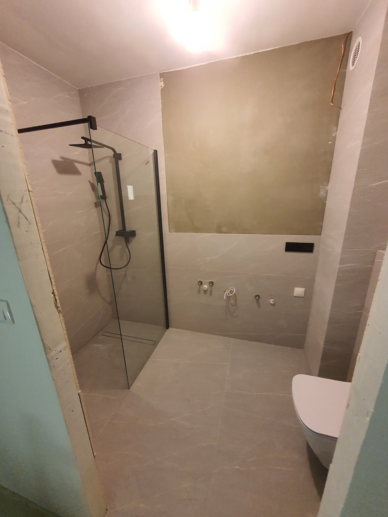 Wnętrze łazienki w trakcie wykańczania, z zamontowaną kabiną prysznicową z czarnymi profilami, częściowo wykończonymi ścianami w odcieniach szarości oraz podwieszaną toaletą