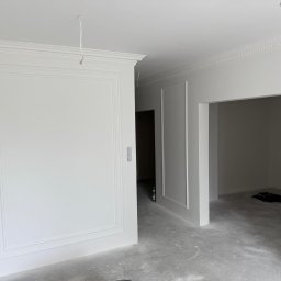 Mat-bud Mateusz Paurowski - Białe ściany z dekoracyjnymi listwami w nowym mieszkaniu, widoczne puszki z farbą, surowa podłoga. Wykończenie wnętrz w minimalistycznym stylu.