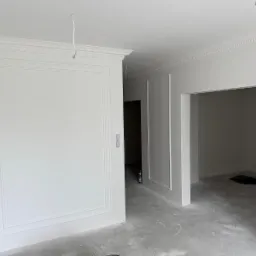 Białe ściany z dekoracyjnymi listwami w nowym mieszkaniu, widoczne puszki z farbą, surowa podłoga. Wykończenie wnętrz w minimalistycznym stylu.