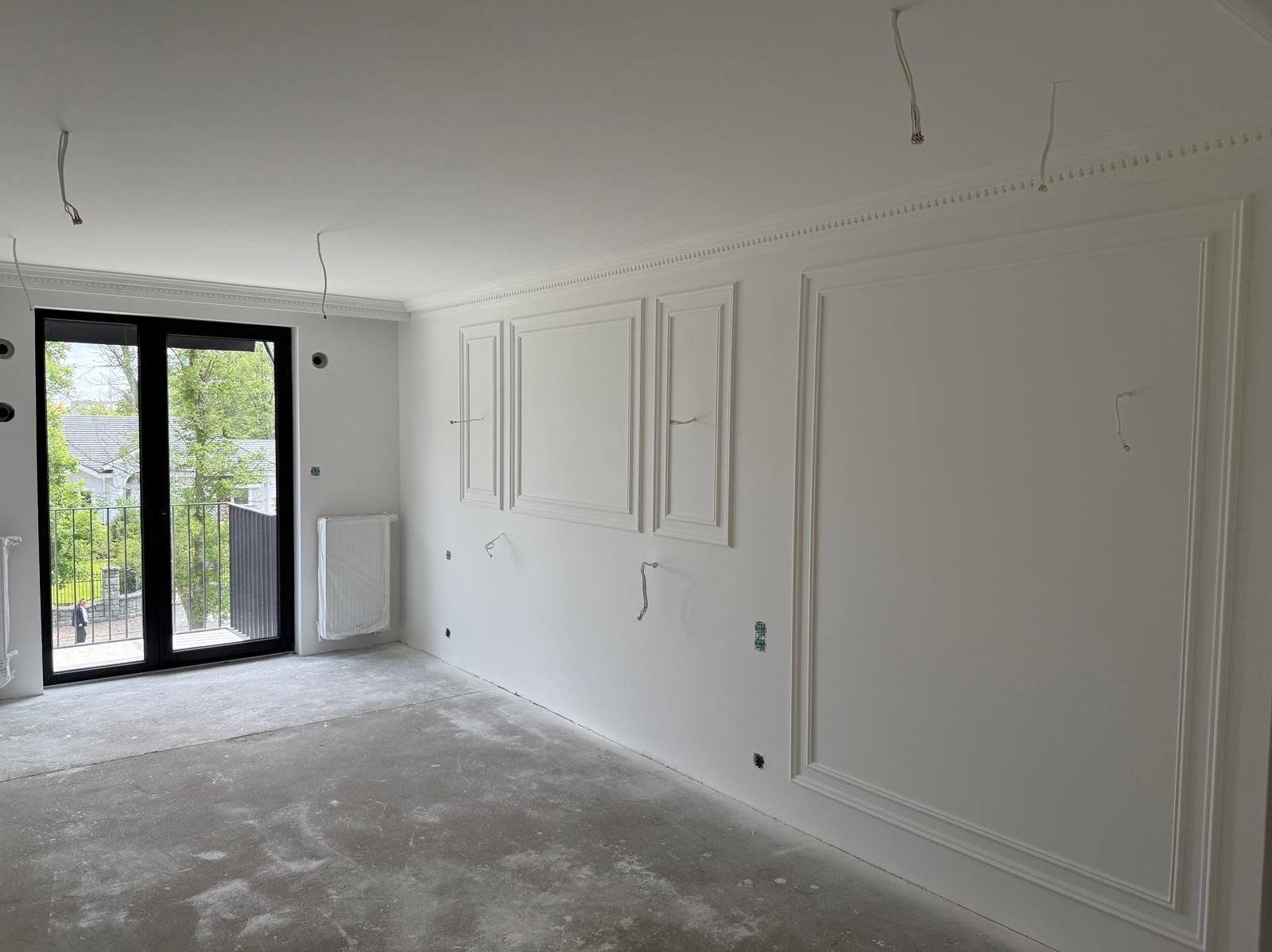 Białe ściany z dekoracyjnymi listwami w nowym wnętrzu, surowy beton na podłodze, okno balkonowe z widokiem na zieleń. Przygotowanie do wykończenia.