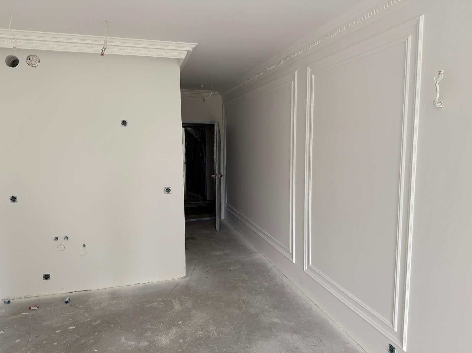 Białe ściany z dekoracyjnymi listwami i sztukaterią w nowym mieszkaniu. Widoczne puszki elektryczne i surowa podłoga. W tle otwarte drzwi.
