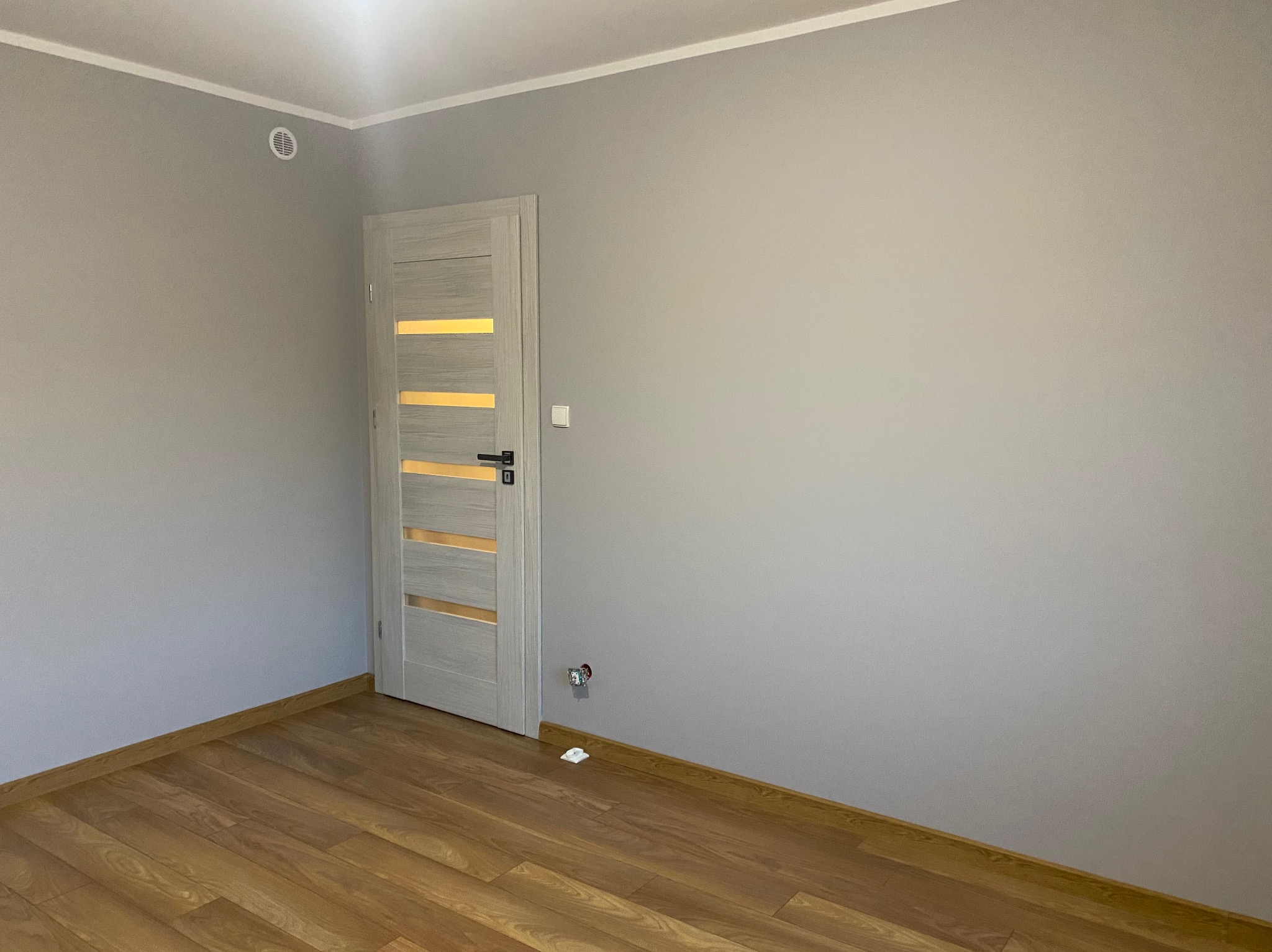 Pokój po remoncie: szare ściany, drewnopodobna podłoga, białe listwy przypodłogowe i drzwi z przeszkleniami. Widoczna puszka elektryczna i wentylacja sufitowa.