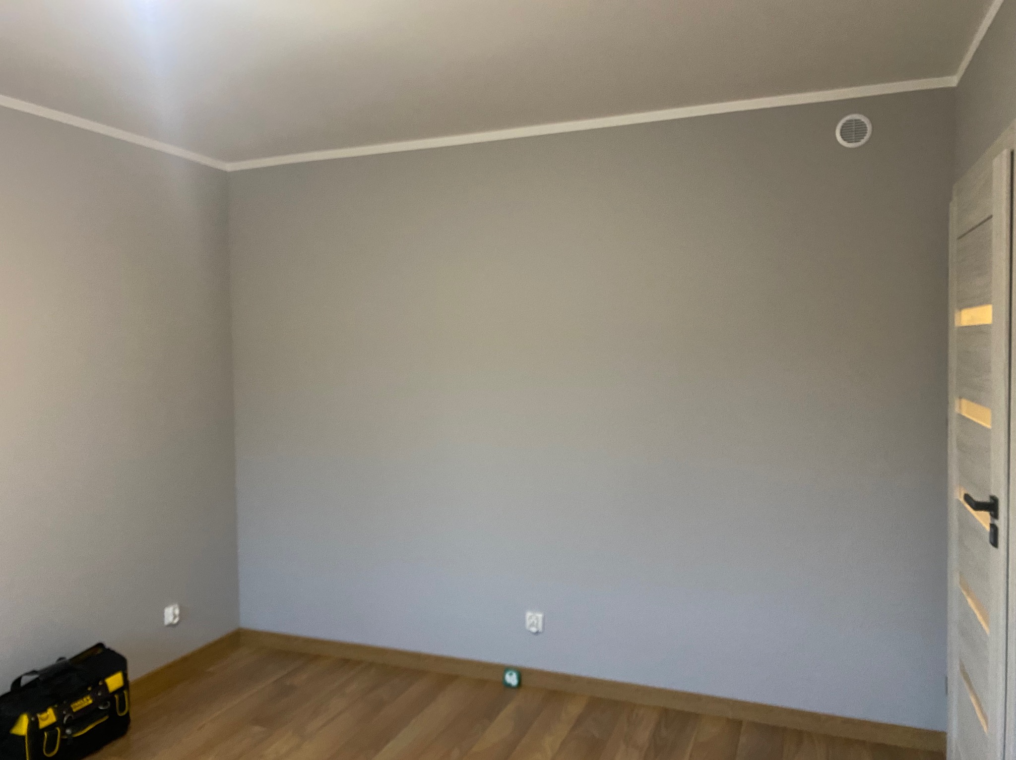 Pokój po malowaniu na szaro, z drewnianą podłogą i białymi listwami. W rogu widoczna skrzynka narzędziowa Stanley i kratka wentylacyjna na ścianie.