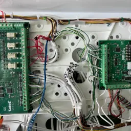 Centrala alarmowa z dwoma modułami elektronicznymi Satel INT-PP v1.4 i Satel INT-E v1.5, widoczne liczne przewody różnej grubości i koloru, umieszczone w białej obudowie ze śrubami.