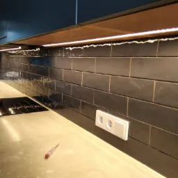 Ciemne, prostokątne płytki na ścianie w kuchni z podświetleniem LED pod szafkami, widoczny blat z płytą indukcyjną i gniazdko elektryczne z testerem napięcia obok.