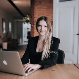 Kobieta w czarnej marynarce siedzi za biurkiem z laptopem, uśmiechając się do kamery w jasnym wnętrzu z otwartymi białymi drzwiami w tle.
