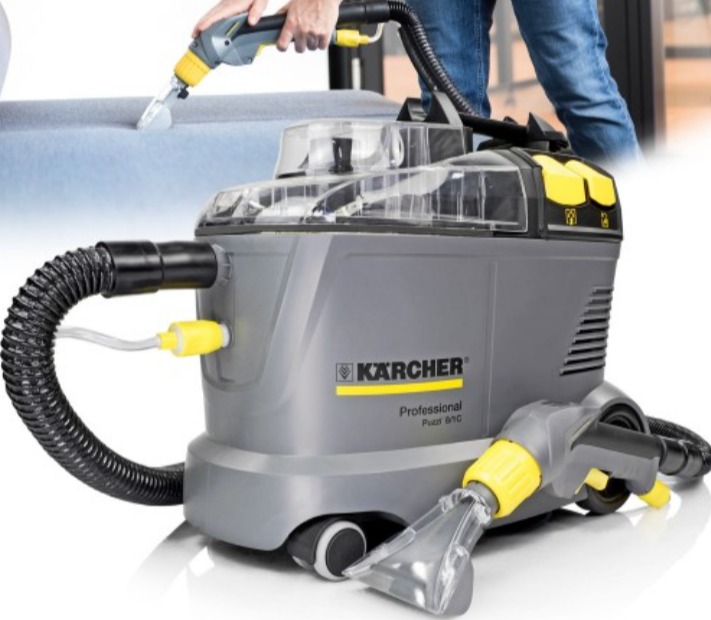Szaro-żółty odkurzacz piorący Kärcher Professional Puzzi 8/1 C w trakcie czyszczenia jasnoniebieskiej kanapy; widoczne dysze i węże, osoba w jeansach w tle.
