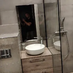 Nowoczesna łazienka po remoncie z kabiną prysznicową typu walk-in, umywalką nablatową na szafce z dwoma szufladami i lustrem, w którym odbija się osoba robiąca zdjęcie.